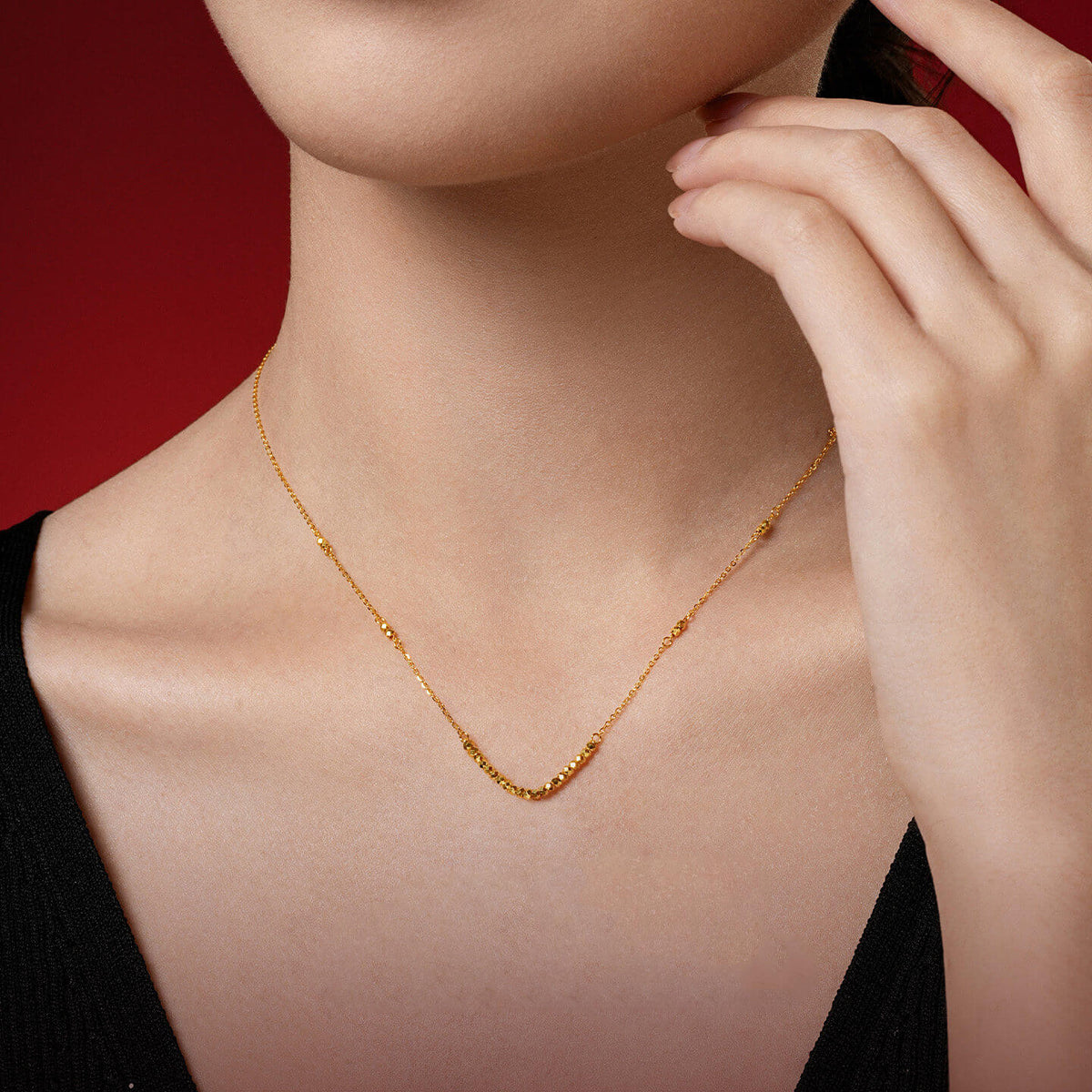 Side view of woman wearing 24K gold disco ball beaded necklace on red background — delicate gold bead chain against black top, elegant fine jewelry styling