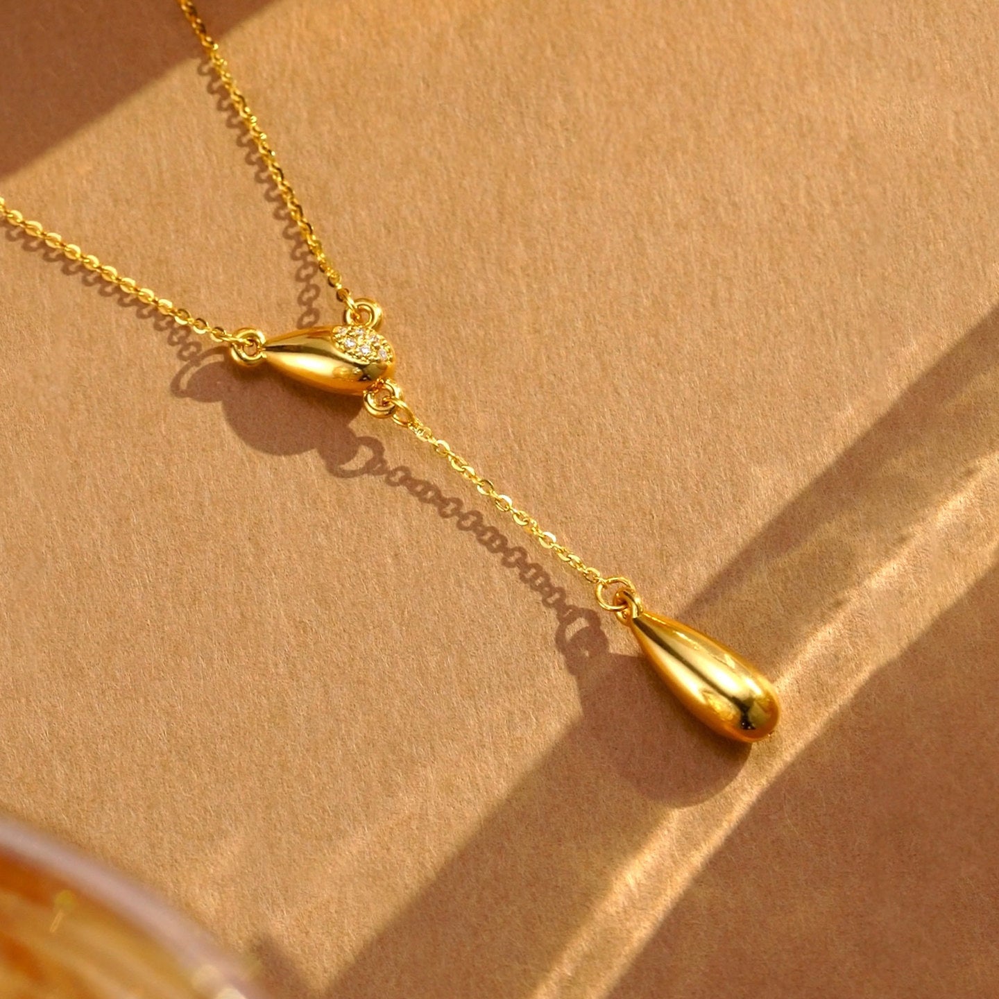 ChowTaiSeng 24K gold teardrop Y necklace styled on kraft paper surface in warm natural sunlight — upper diamond-inlaid teardrop and round diamond cluster catching light, tassel chain casting delicate shadow, larger polished gold teardrop drop below