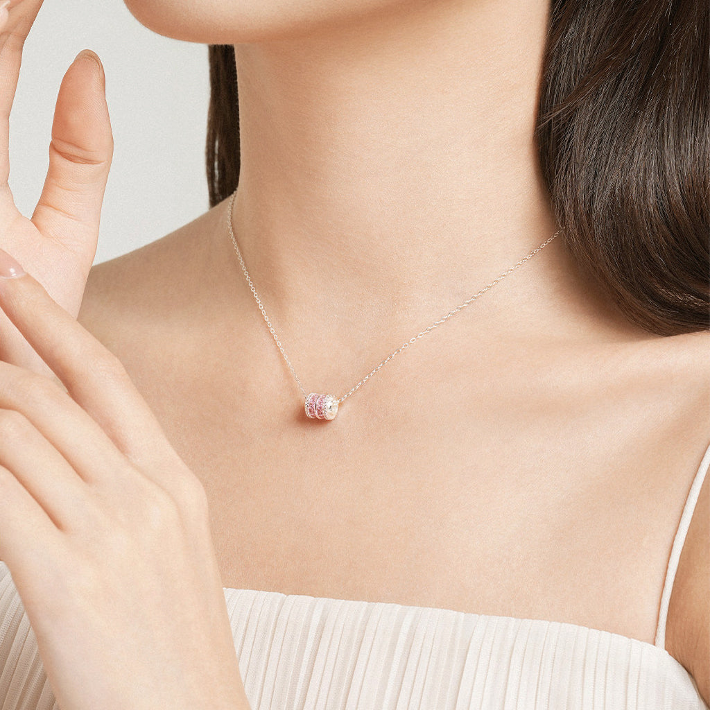 Close-up of Asian model's neck and hand wearing ChowTaiSeng silver pink cubic zirconia barrel pendant necklace on silver cable chain, white strapless top, light grey background