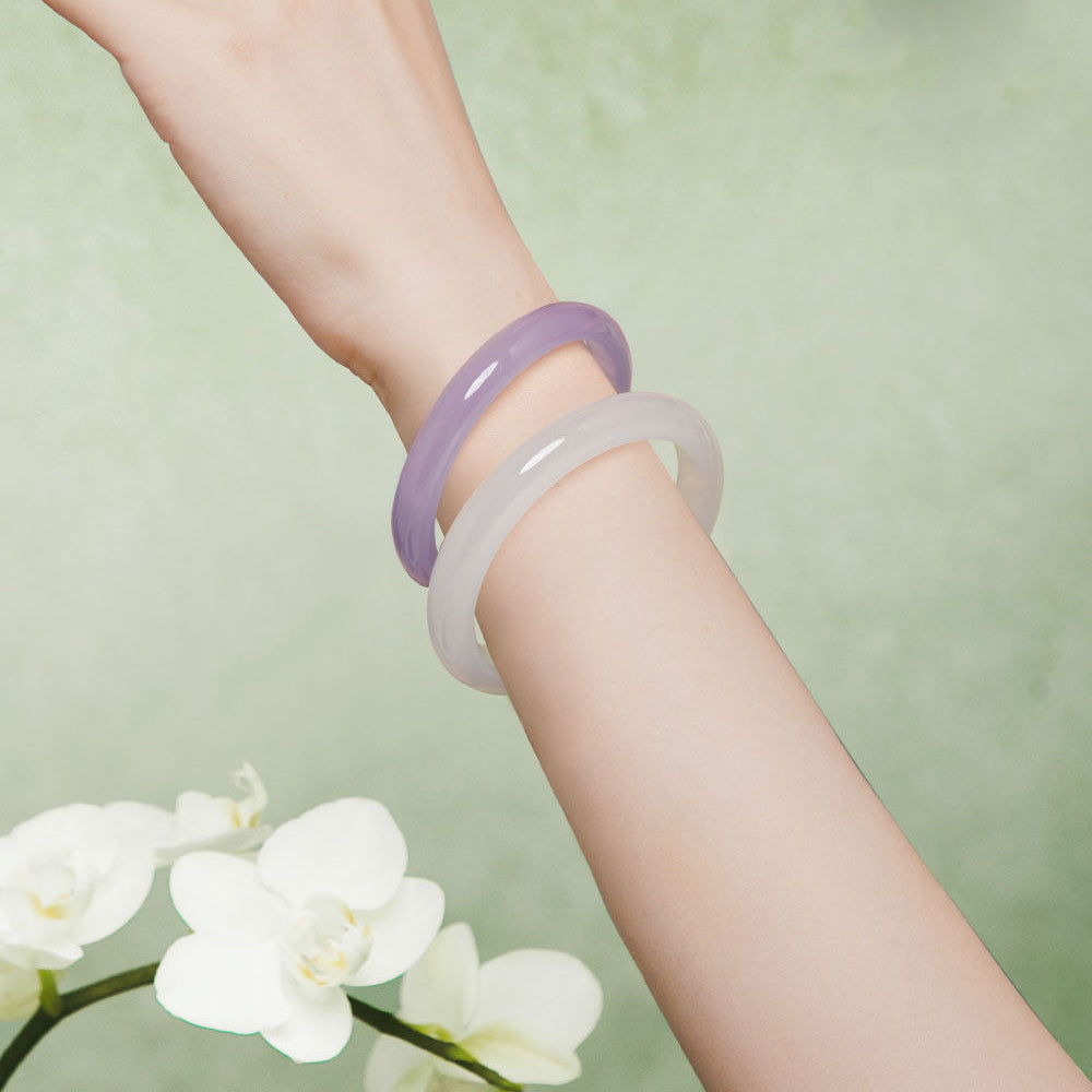 Two smooth translucent bangles, one light purple and one pale frosted white, worn on a person's wrist against a soft green background with white flowers at the lower left.
