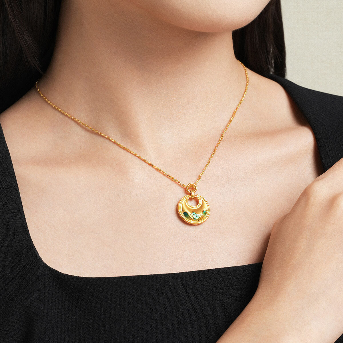 Model wearing ChowTaiSeng 24K Gold Round Ruyi Lock Pendant with green enamel mountain motif, black top, close-up neckline shot