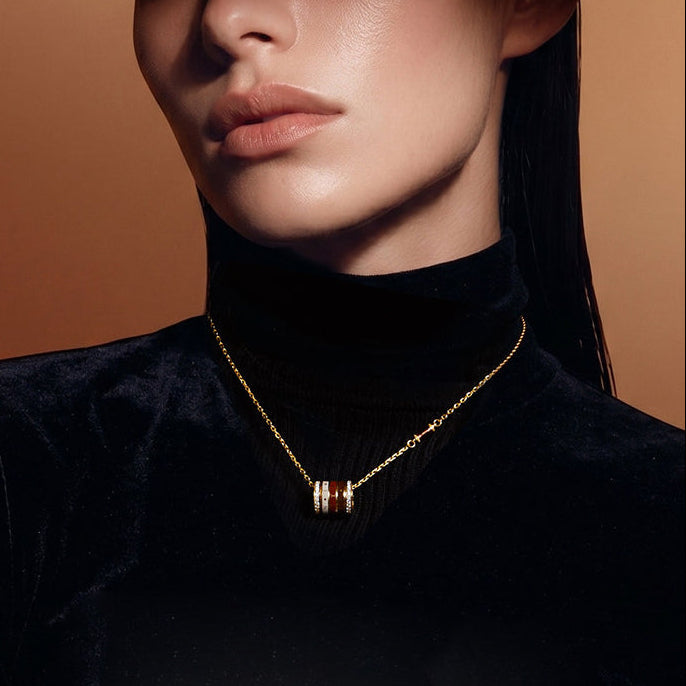 A close-up of a model wearing a delicate gold chain necklace with a cylindrical pendant featuring red and white striped accents, styled with a black velvet turtleneck against a warm brown background.