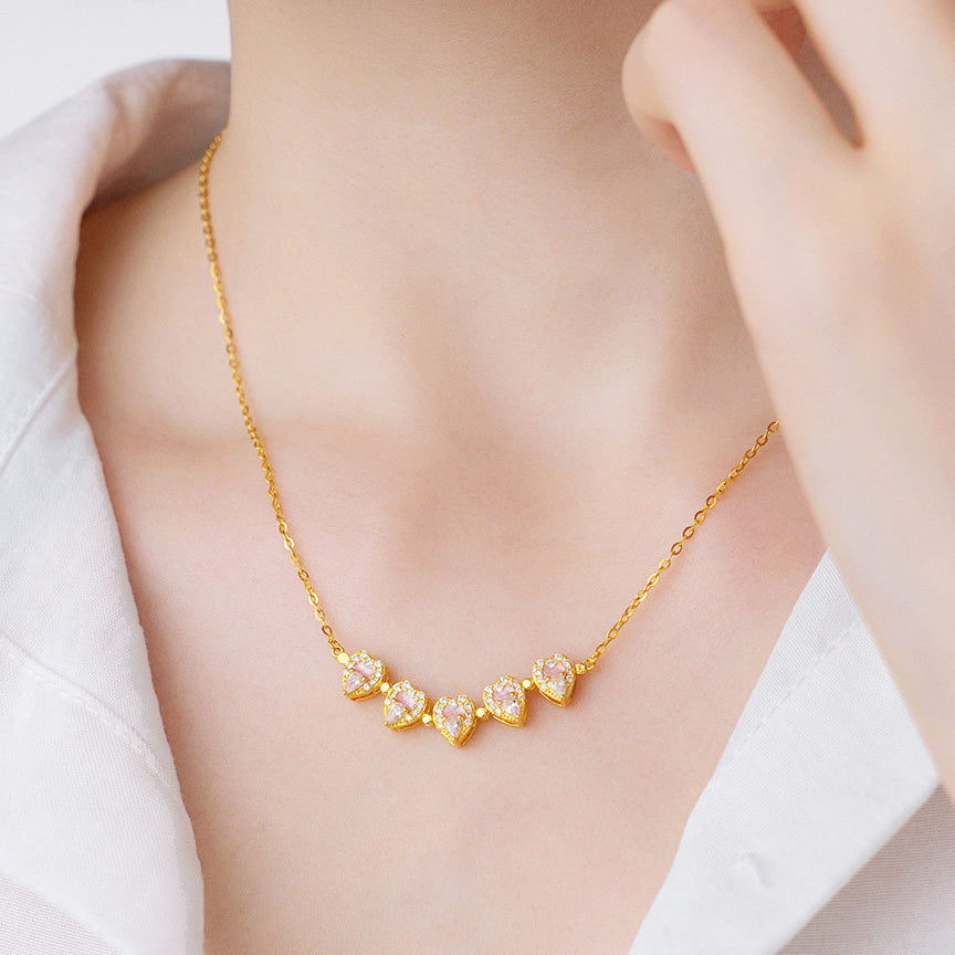 Model wearing ChowTaiSeng gold plated silver peach blossom heart necklace in assembled flower mode — five pink enamel and CZ heart pendants forming a full peach blossom pendant at neckline on gold cable chain, white shirt background