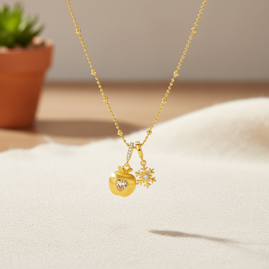 ChowTaiSeng gold plated snowflake apple necklace hanging against warm neutral background with terracotta plant, gold beaded chain with apple and snowflake dual charms